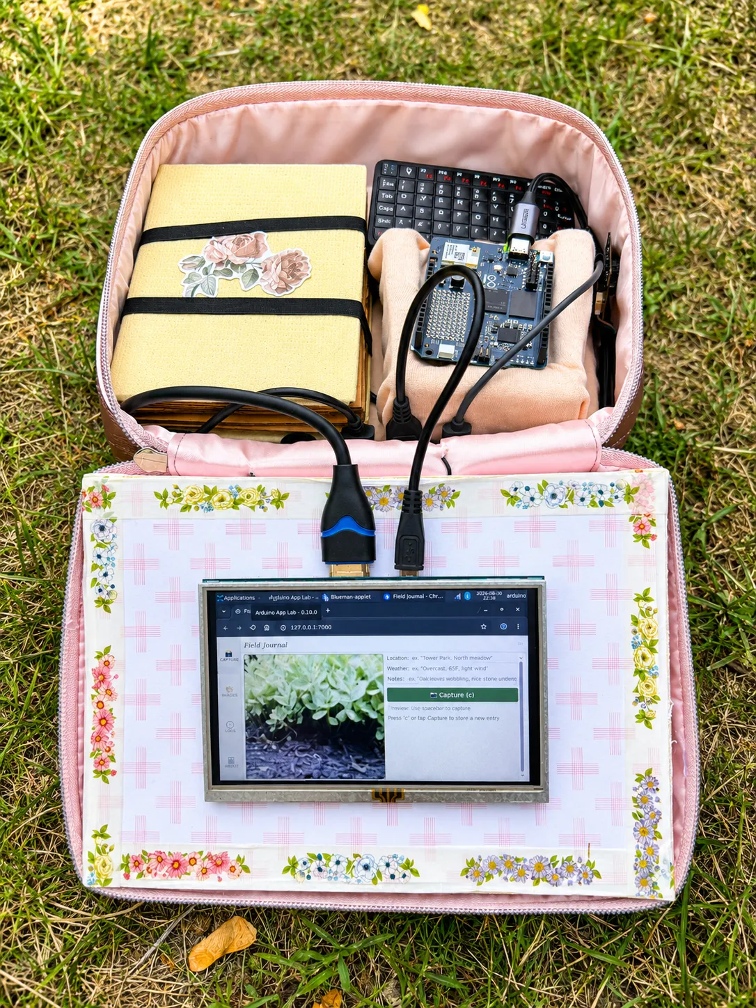 A floral zippered case open outdoors near green leaves, holding a small touchscreen, a keyboard, and a wired Arduino board tucked into fabric pockets.