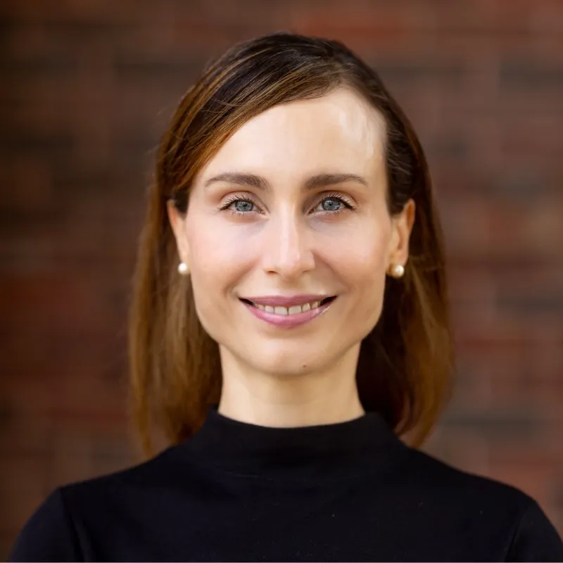 Francesca Lazzeri smiling in front of a brick wall, wearing a black turtleneck and pearl earrings.