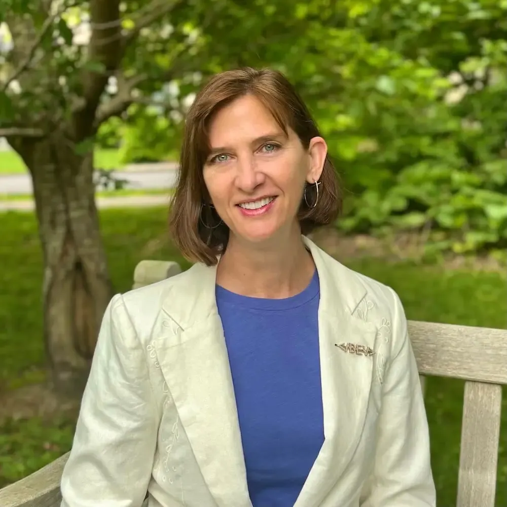 Jen Looper smiling outdoors on a wooden bench, wearing a cream blazer over a blue top, with trees in the background.