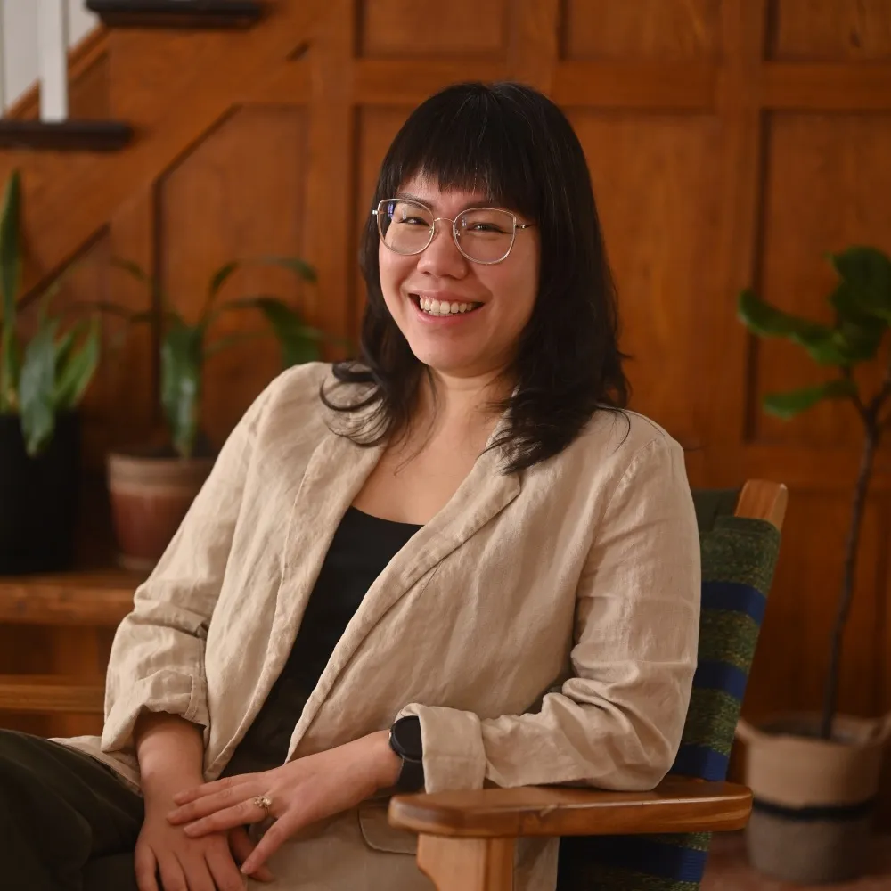 Jess Lee smiling in a wood-paneled room, wearing glasses and a tan blazer, seated in a wooden chair.