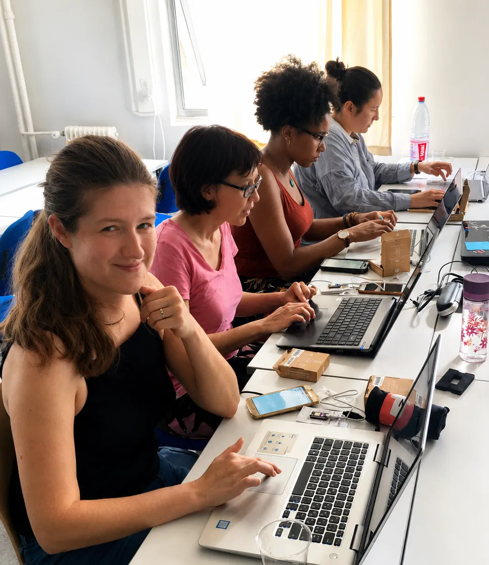 Four women sit side by side at a long table, laptops open, wiring up hardware kits together during a workshop session.